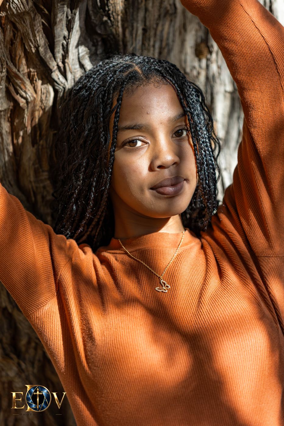 Close portrait of a young woman smiling, arms raised against a tree trunk